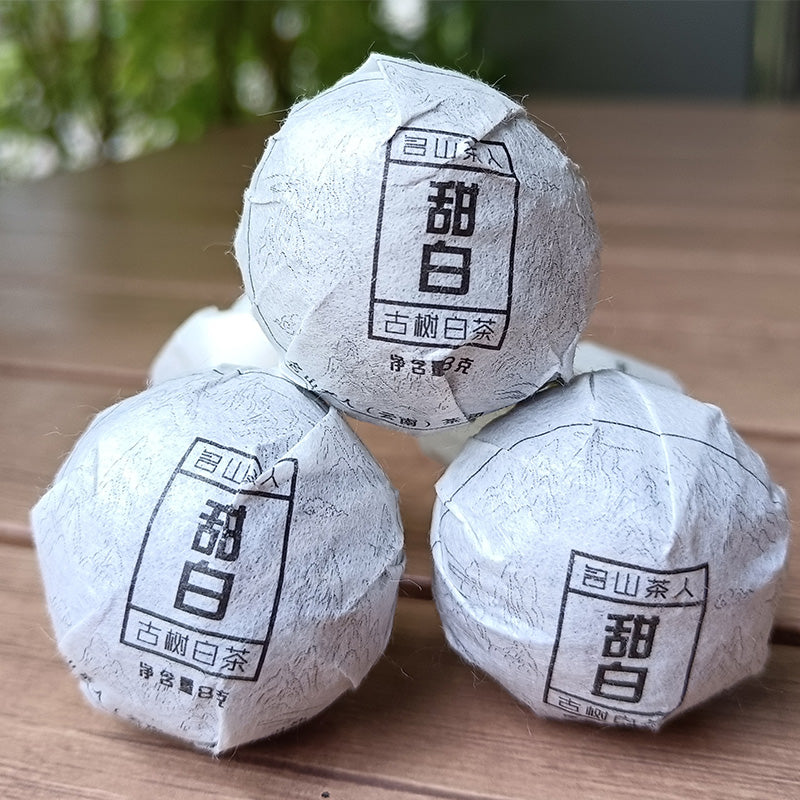 Three Sweet White Antique Tree Beads 8g/80g wrapped in thin paper with Chinese characters, arranged in a pyramid shape, sit on a wooden surface with a blurred greenery background, showcasing the rustic charm of ZenTea Manor-YN001.