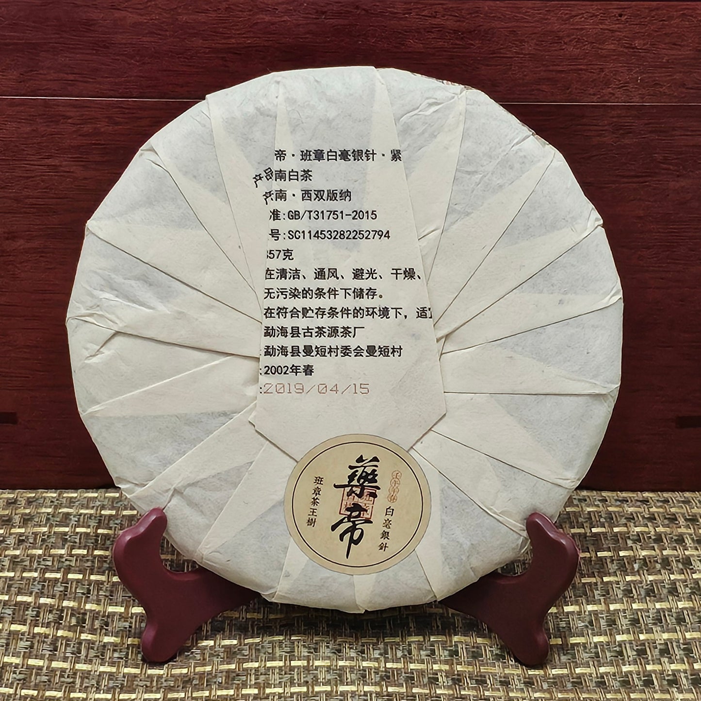 Wrapped round package of 2002 Banzhang ( 班章 ) Tea King Ancient Tree ( 茶王古樹 ) White Hair Silver Needle White Tea ( 白毫銀針白茶 ) 357g by ZenTea Manor-YN003, featuring text in Chinese and English on its front. The packaging is displayed upright on a wooden stand against a wooden background. Banzhang Ancient Tree tea leaves add an authentic touch to this exquisite offering.