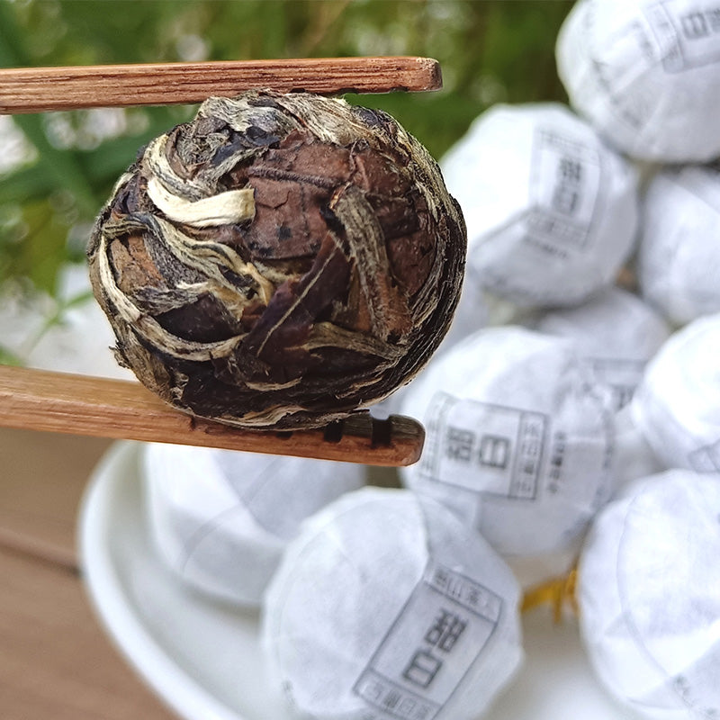 A close-up of chopsticks holding a ball of compressed tea leaves, hinting at the wild flavor of Sweet White Antique Tree Beads 8g/80g by ZenTea Manor-YN001, with more wrapped tea balls in the background.