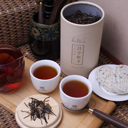 A canister of tea, two cups of poured tea, a tea pitcher, loose tea leaves, and round biscuits are arranged neatly on a wooden tray on a woven surface.