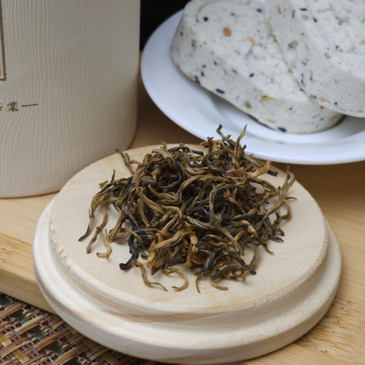 A small pile of loose tea leaves sits on a round wooden lid. In the background, a white plate holds a couple of rice cakes.