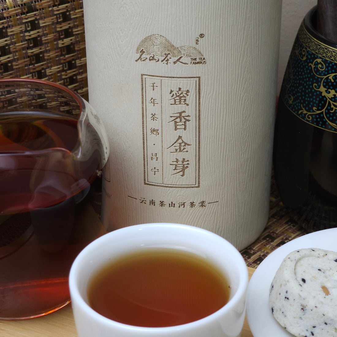 A glass teapot was filled with tea, a white teacup was filled with tea, and there was a decorative tin container with Chinese characters written on it on the table. Small desserts or snacks can be seen on the right side.