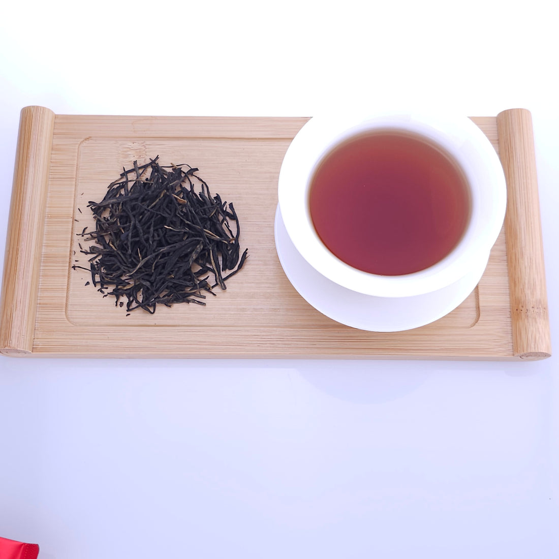 A white cup filled with brown tea sits on a wooden tray next to a small pile of dried tea leaves.