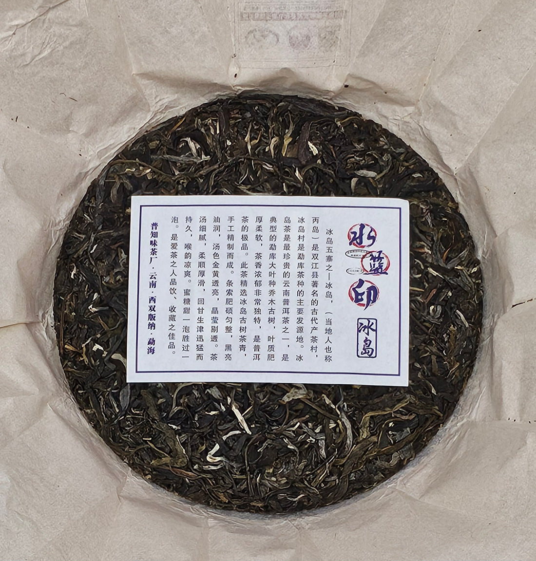 A round cake of compressed tea leaves wrapped in paper. The center features a rectangular label with text in Chinese characters, indicating its origin from Bingdao Old Village.