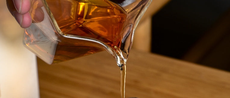 Pour the amber liquid from the glass decanter into a container on a wooden surface.