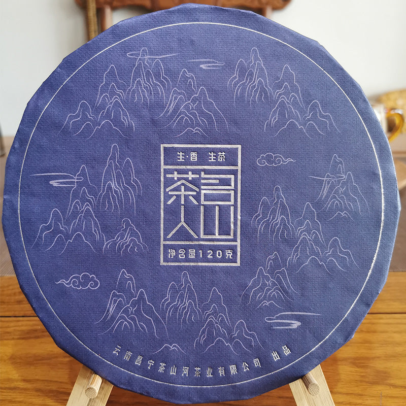 A round, blue-wrapped tea cake featuring intricate mountain designs and Chinese characters is displayed on a wooden stand, showcasing traditional craft refined in the heart of Yunnan high mountain tea regions. Product Name: Rooibos Tea 120g Brand Name: Zen Tea Master