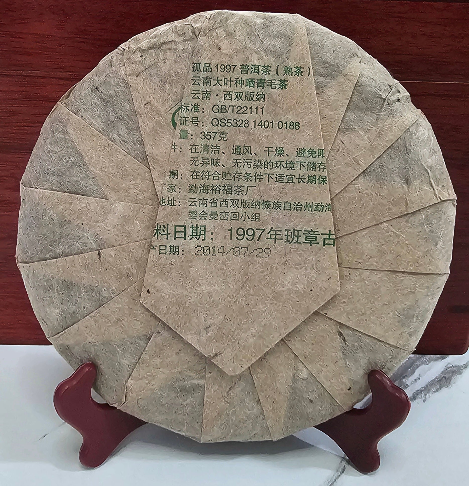 A round, packaged tea cake with Chinese text on the wrapper, resting on a wooden display stand with a reddish-brown backdrop. The packaging reveals it as 1997 Years Banzhang ( 班章 ) Ancient Tree Puerh Ripe Tea 357g by ZenTea Manor-YN002, promising the unique flavors of ancient tree tea.