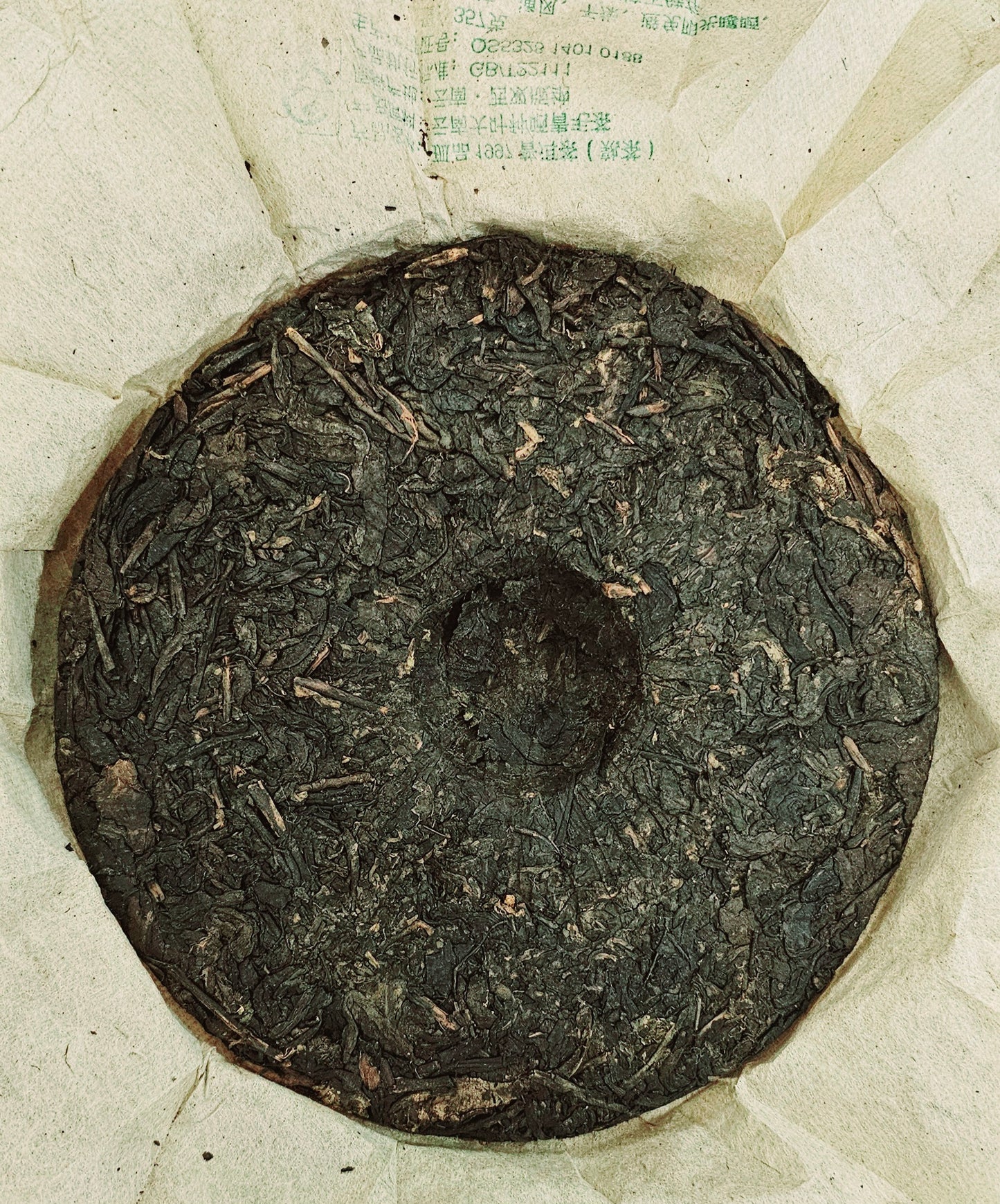 A round block of ancient tree tea sits on beige paper with some printed text visible near the edges, hinting at its storied origins. This 1997 Years Banzhang (班章) Ancient Tree Puerh Ripe Tea 357g radiates a rich, dark essence, capturing the timeless allure of ZenTea Manor-YN002.