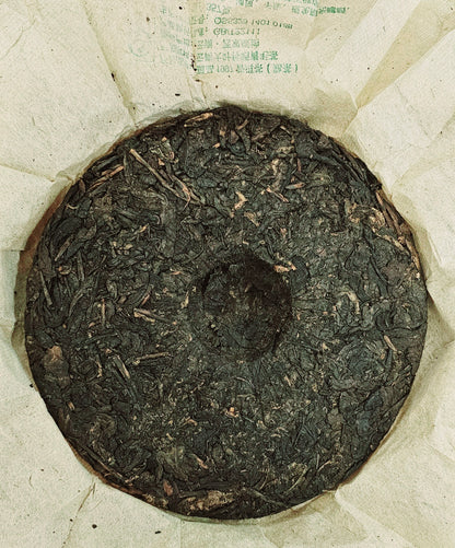 A round block of ancient tree tea sits on beige paper with some printed text visible near the edges, hinting at its storied origins. This 1997 Years Banzhang (班章) Ancient Tree Puerh Ripe Tea 357g radiates a rich, dark essence, capturing the timeless allure of ZenTea Manor-YN002.