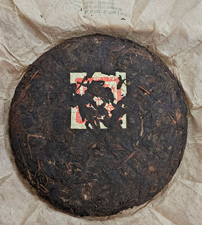 A round disc of dark, compressed 1997 Years Banzhang ( 班章 ) Ancient Tree Puerh Ripe Tea 357g from ZenTea Manor-YN002 with a paper label partially visible in the center, resting on crumpled brown parchment paper.