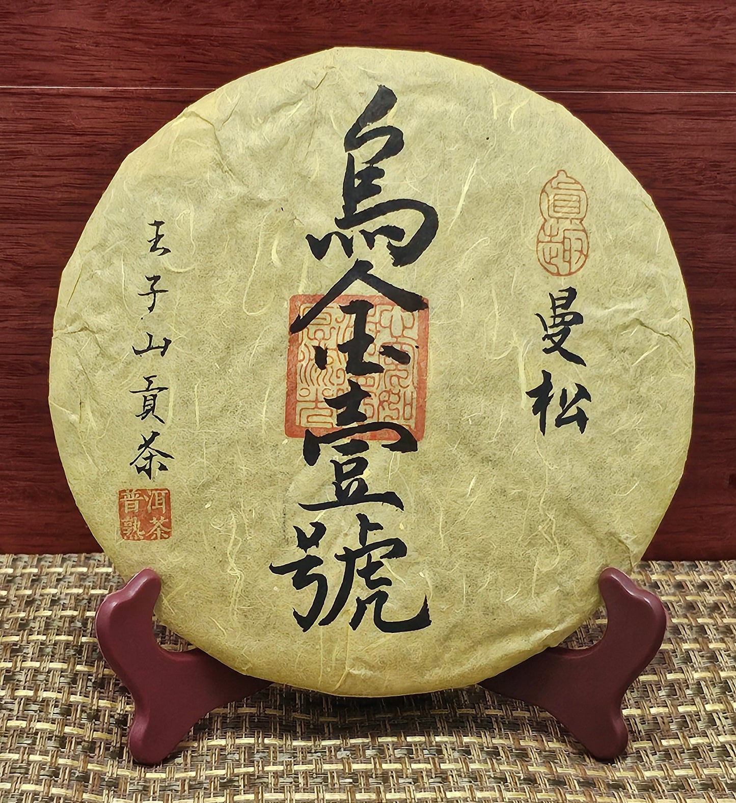 A round package of 2008 Mansong Prince Mountain Tribute Tea ( 曼松王子山貢茶 ) - Wujin No.1 ( 烏金壹號 ) Puerh Ripe Tea 357g from ZenTea Manor-YN005 with Chinese characters on the front sits on a wooden stand atop a woven mat, with a wood-paneled background.