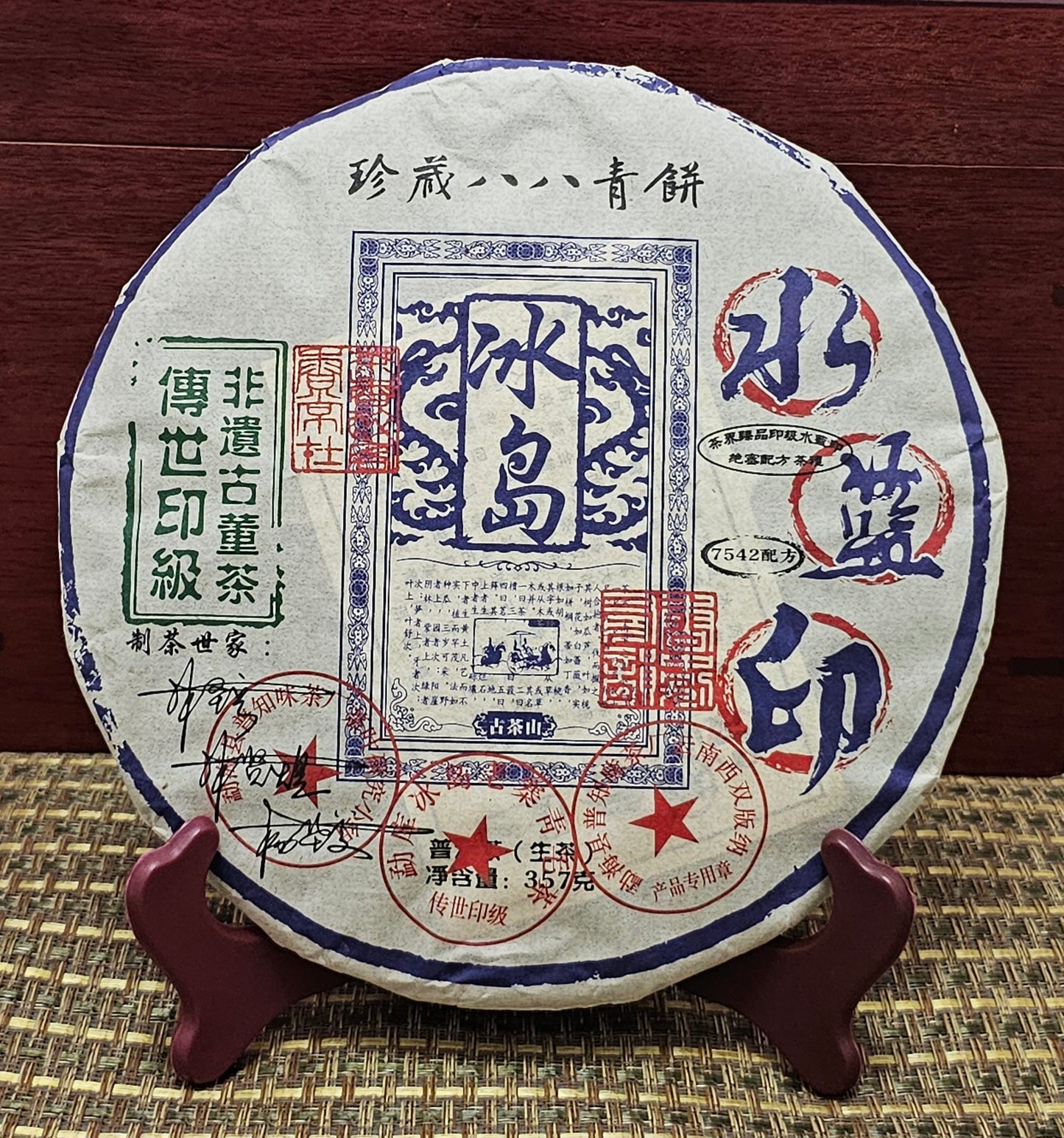 2016 Bingdao Old Village (冰島老寨) - Water Blue Seal 88 Green Cakes (水藍印八八青餅) Pu'er Raw Tea by ZenTea Manor-YN005, featuring stamps and Chinese text, displayed upright on a wooden stand against a woven mat background.