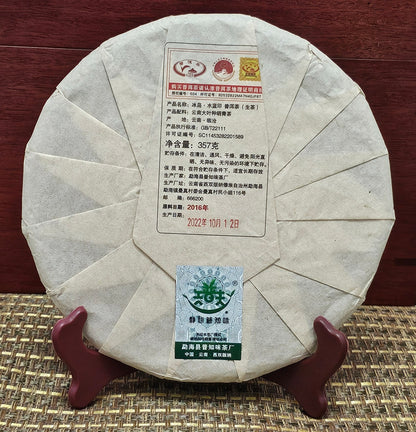 A round, beige-wrapped tea cake labeled with Chinese text, rests on a wooden stand atop a woven mat. The label includes red stamps and production details of the "2016 Bingdao Old Village ( 冰島老寨 ) - Water Blue Seal 88 Green Cakes ( 水藍印八八青餅 ) Puerh Raw Tea 357g" from ZenTea Manor-YN005.