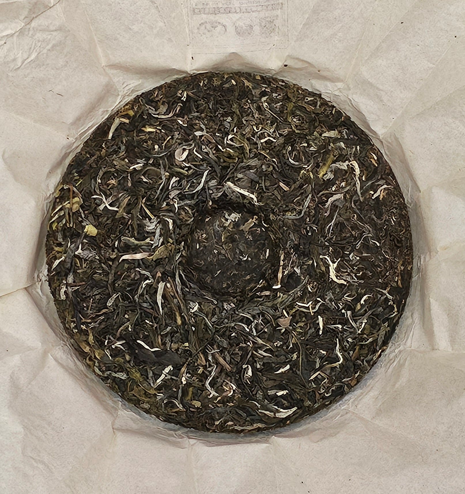 A round cake of dried tea leaves, wrapped in thin, white paper, known as the 2016 Bingdao Old Village ( 冰島老寨 ) - Water Blue Seal 88 Green Cakes ( 水藍印八八青餅 ) Puerh Raw Tea 357g by ZenTea Manor-YN005.