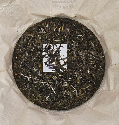 A circular compressed tea cake made of loose tea leaves, labeled as the 2016 Bingdao Old Village ( 冰島老寨 ) - Water Blue Seal 88 Green Cakes ( 水藍印八八青餅 ) Puerh Raw Tea 357g from ZenTea Manor-YN005, is centered on a piece of crumpled white parchment paper.