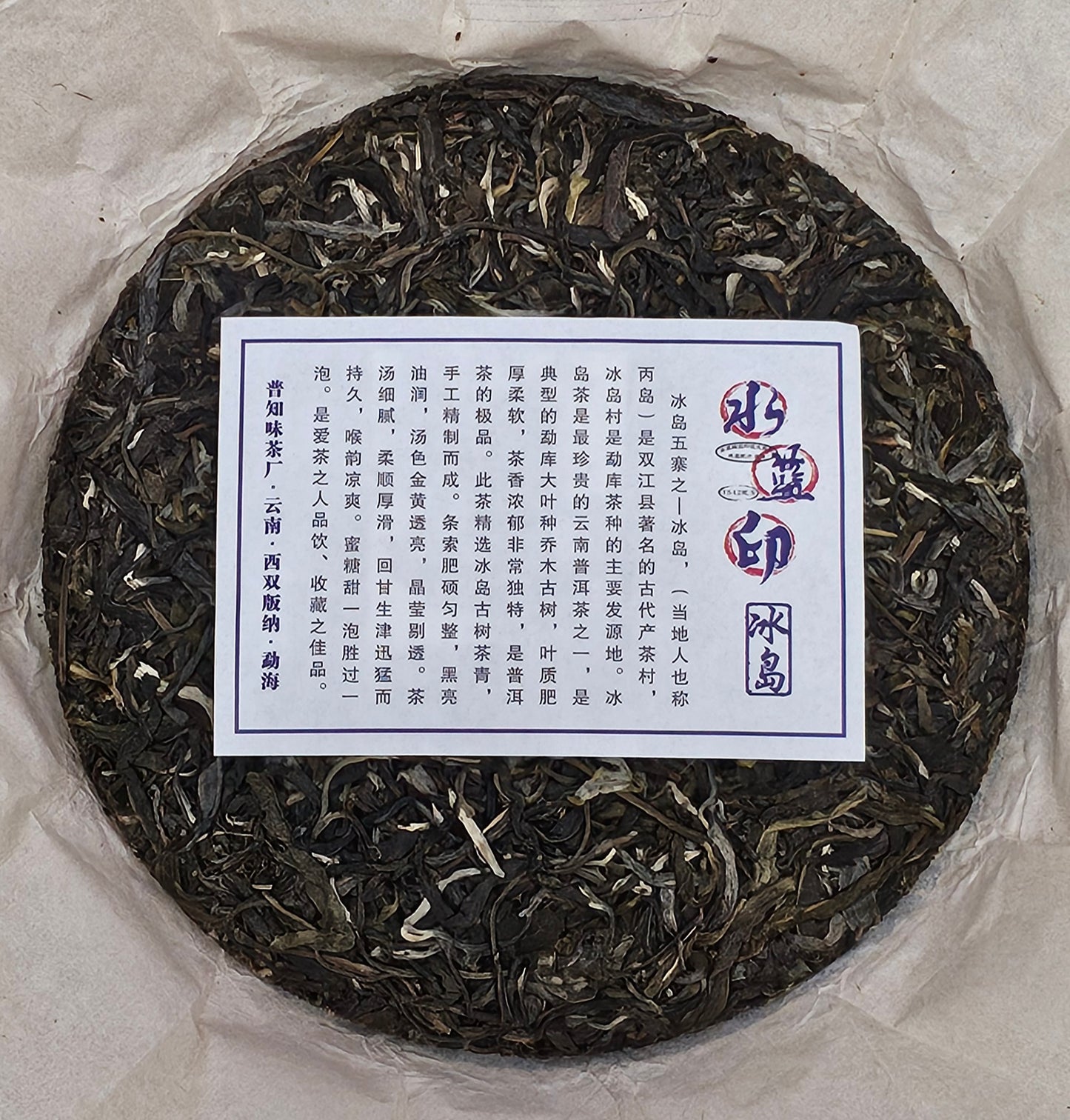 A round compressed tea cake, the 2016 Bingdao Old Village (冰島老寨) - Water Blue Seal 88 Green Cakes (水藍印八八青餅), with a label featuring Chinese text at the center. The tea leaves in the 357g Puerh Raw Tea cake by ZenTea Manor-YN005 appear dark and loosely compacted.