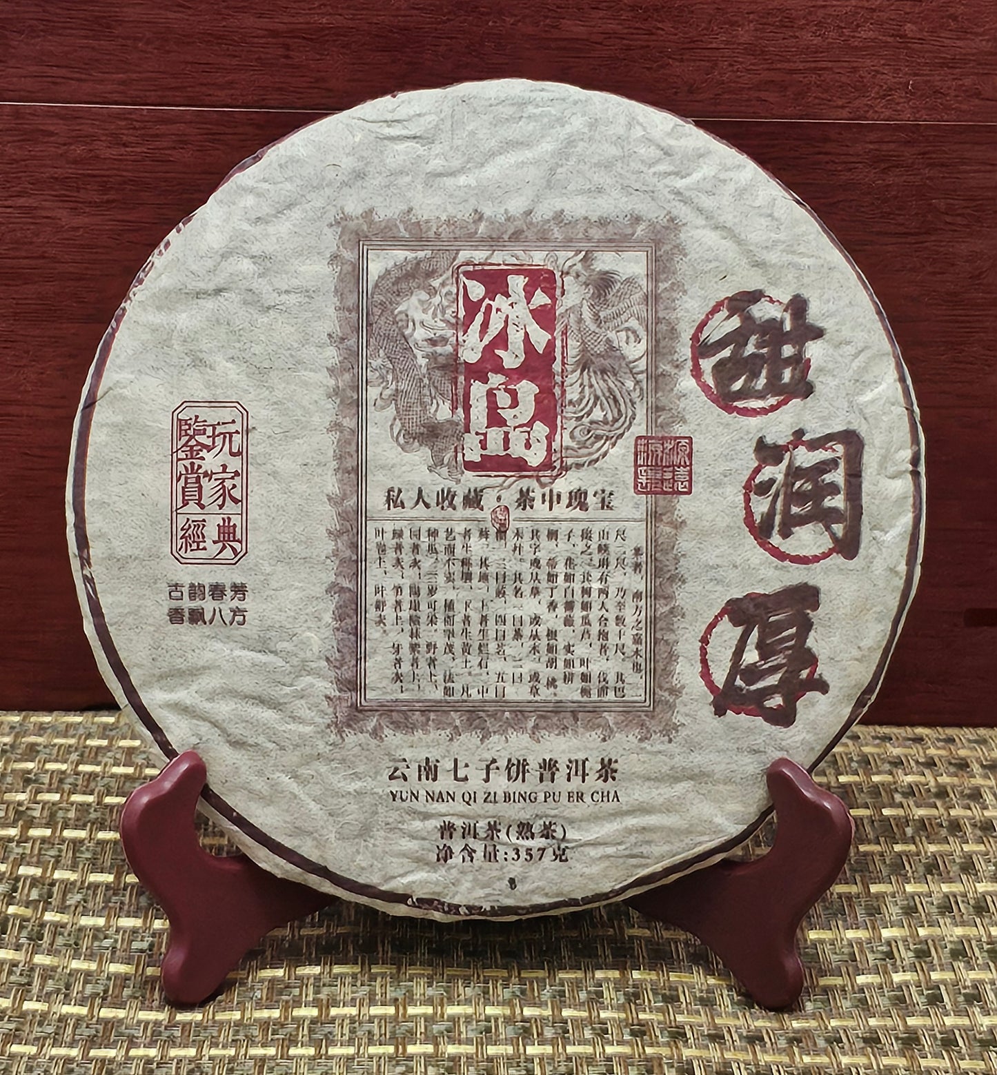 A round, traditionally wrapped tea cake labeled "2005 Bingdao ( 冰島 )• Sweet and Mellow Thick ( 甜潤厚 ) Puerh Ripe Tea 357g" from ZenTea Manor-YN005, displayed on a wooden stand against a red wooden background. Chinese characters and vintage design details enhance the packaging of this aged tea.