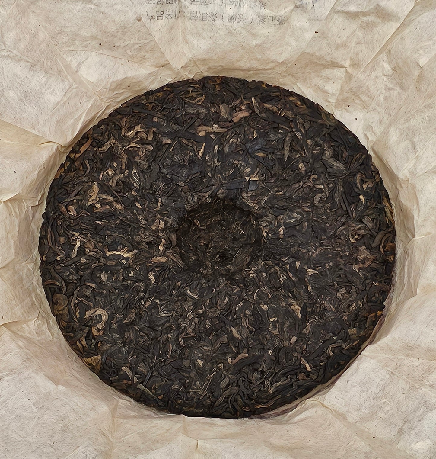 A circular block of compressed 2005 Bingdao ( 冰島 )• Sweet and Mellow Thick ( 甜潤厚 ) Puerh Ripe Tea 357g with a hole in the middle, resting on a piece of crumpled parchment paper from ZenTea Manor-YN005.