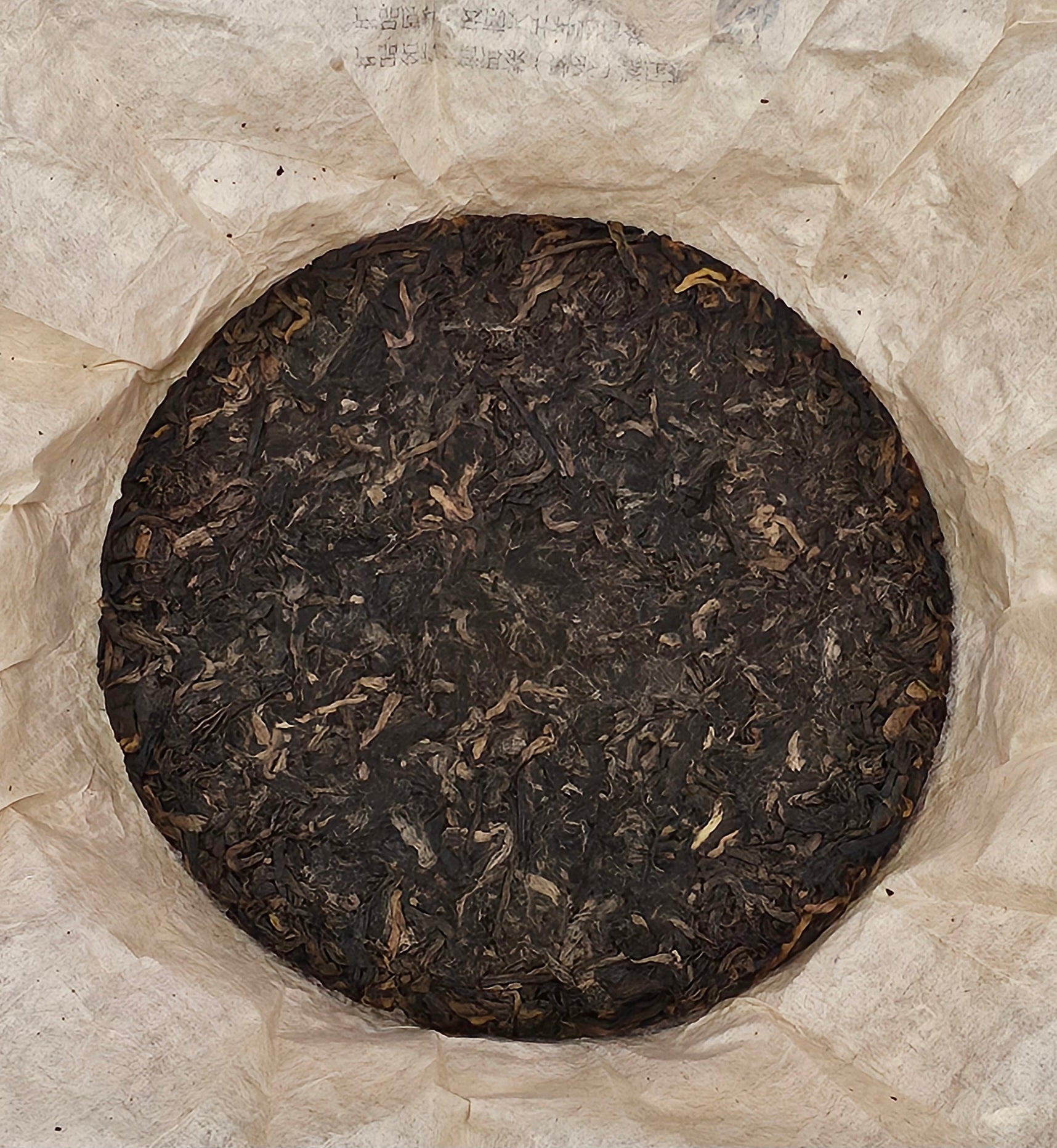 A round, compressed disk of 2005 Bingdao (冰島)• Sweet and Mellow Thick (甜潤厚) Puerh Ripe Tea 357g from ZenTea Manor-YN005 is shown wrapped in white crinkled paper.