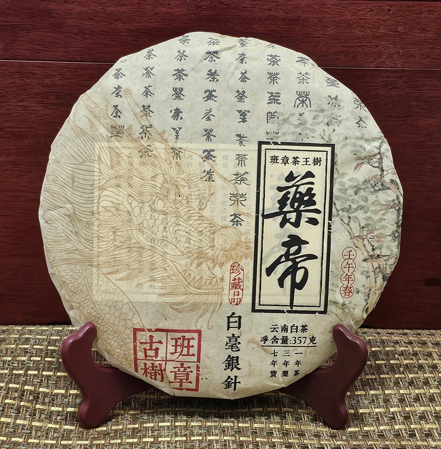 A round, wrapped package of 2002 Banzhang (班章) Tea King Ancient Tree (茶王古樹) White Hair Silver Needle White Tea (白毫銀針白茶) 357g by ZenTea Manor-YN003 with Chinese text, including red and black characters, and a dragon illustration on the front, placed on a wooden stand against a red wood background.