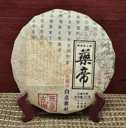 A round, wrapped package of 2002 Banzhang (班章) Tea King Ancient Tree (茶王古樹) White Hair Silver Needle White Tea (白毫銀針白茶) 357g by ZenTea Manor-YN003 with Chinese text, including red and black characters, and a dragon illustration on the front, placed on a wooden stand against a red wood background.