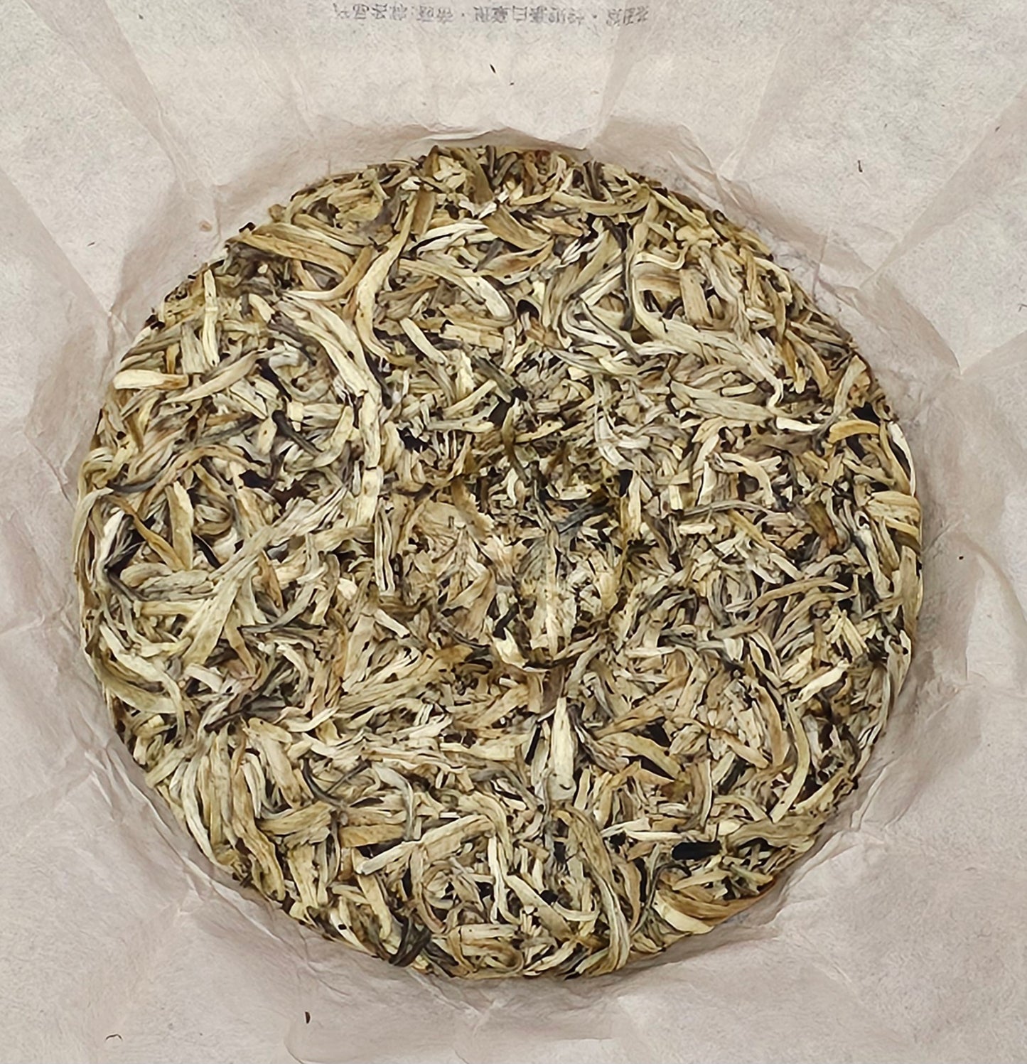 A round cake of white tea enclosed in white parchment paper, showcasing tightly packed, dried tea leaves ranging in color from light beige to dark brown. This exquisite blend includes delicate White Hair Silver Needle leaves, enhancing its refined character and aroma. This is the 2002 Banzhang (班章) Tea King Ancient Tree (茶王古樹) White Hair Silver Needle White Tea (白毫銀針白茶) 357g from ZenTea Manor-YN003.