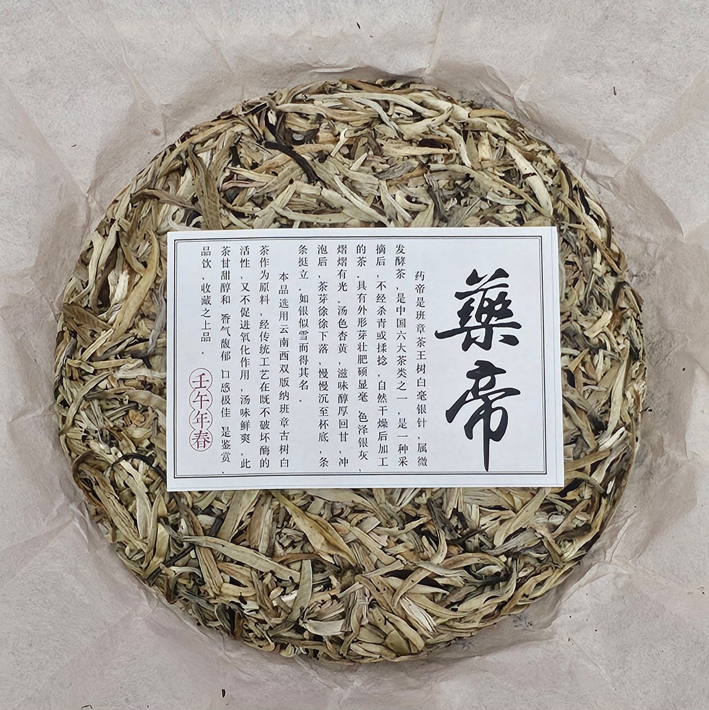 A round block of 2002 Banzhang ( 班章 ) Tea King Ancient Tree ( 茶王古樹 ) White Hair Silver Needle White Tea ( 白毫銀針白茶 ) 357g from ZenTea Manor-YN003 with a rectangular label in Chinese characters on top, placed delicately on parchment paper.