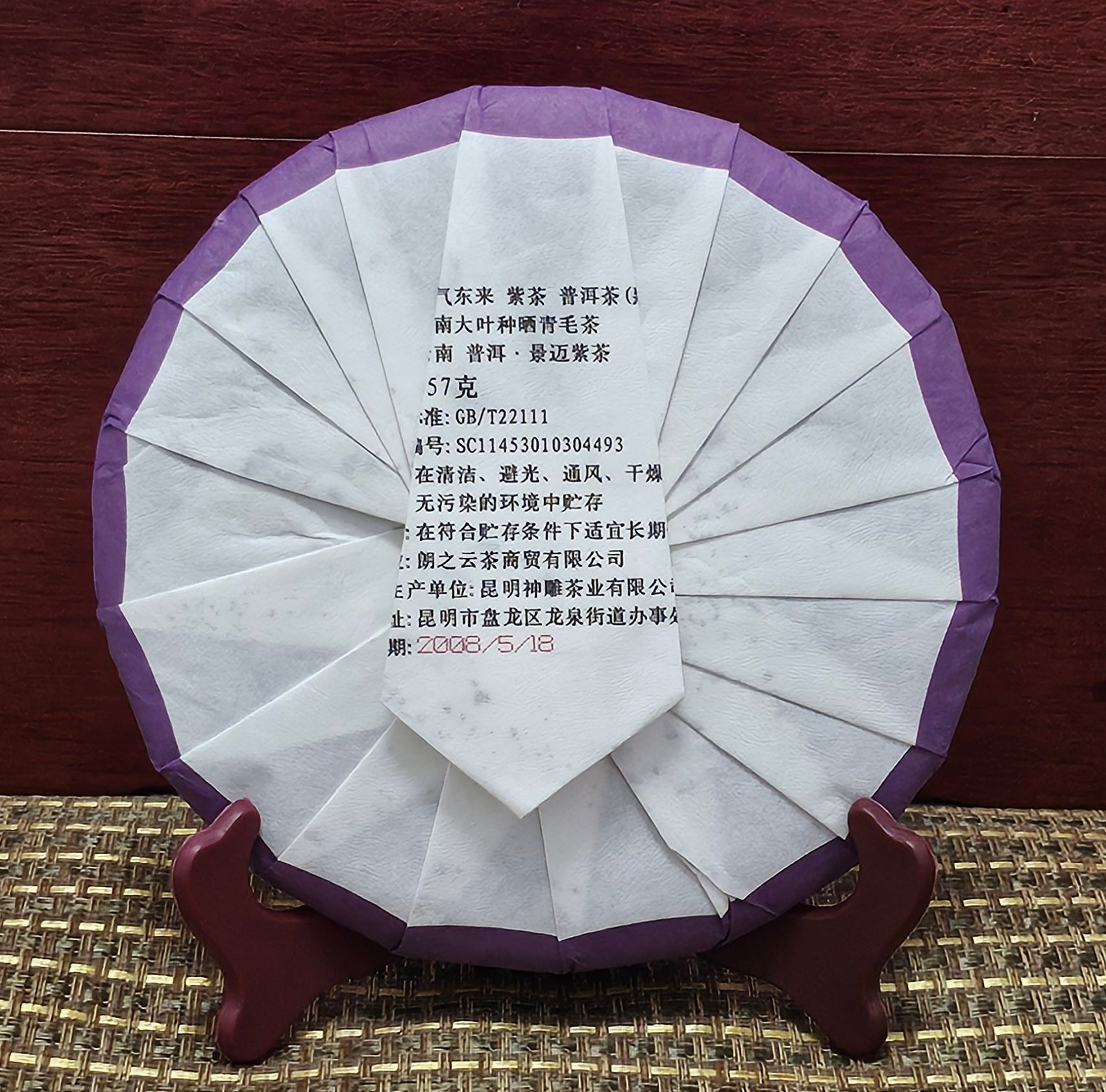 A round, wrapped tea cake of 2008 Jingmai Mountain (景邁山) Purple Tea (紫茶) 357g from ZenTea Manor-YN003 stands on a wooden holder. The white and purple packaging has printed text in Chinese, product details, and a production date of 2008/5/18. Crafted from leaves of ancient tea trees, this Jingmai Purple Tea promises a rich, storied flavor.