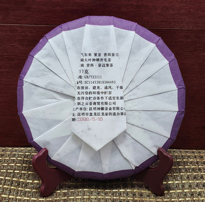 A round, wrapped tea cake of 2008 Jingmai Mountain (景邁山) Purple Tea (紫茶) 357g from ZenTea Manor-YN003 stands on a wooden holder. The white and purple packaging has printed text in Chinese, product details, and a production date of 2008/5/18. Crafted from leaves of ancient tea trees, this Jingmai Purple Tea promises a rich, storied flavor.