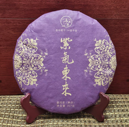 A round, purple package of 2008 Jingmai Mountain ( 景邁山 ) Purple Tea ( 紫茶 ) 357g from ZenTea Manor-YN003 with intricate gold floral designs displayed on a wooden stand. The packaging has Chinese characters and weighs 357 grams, reflecting the rich heritage of ancient tea trees.