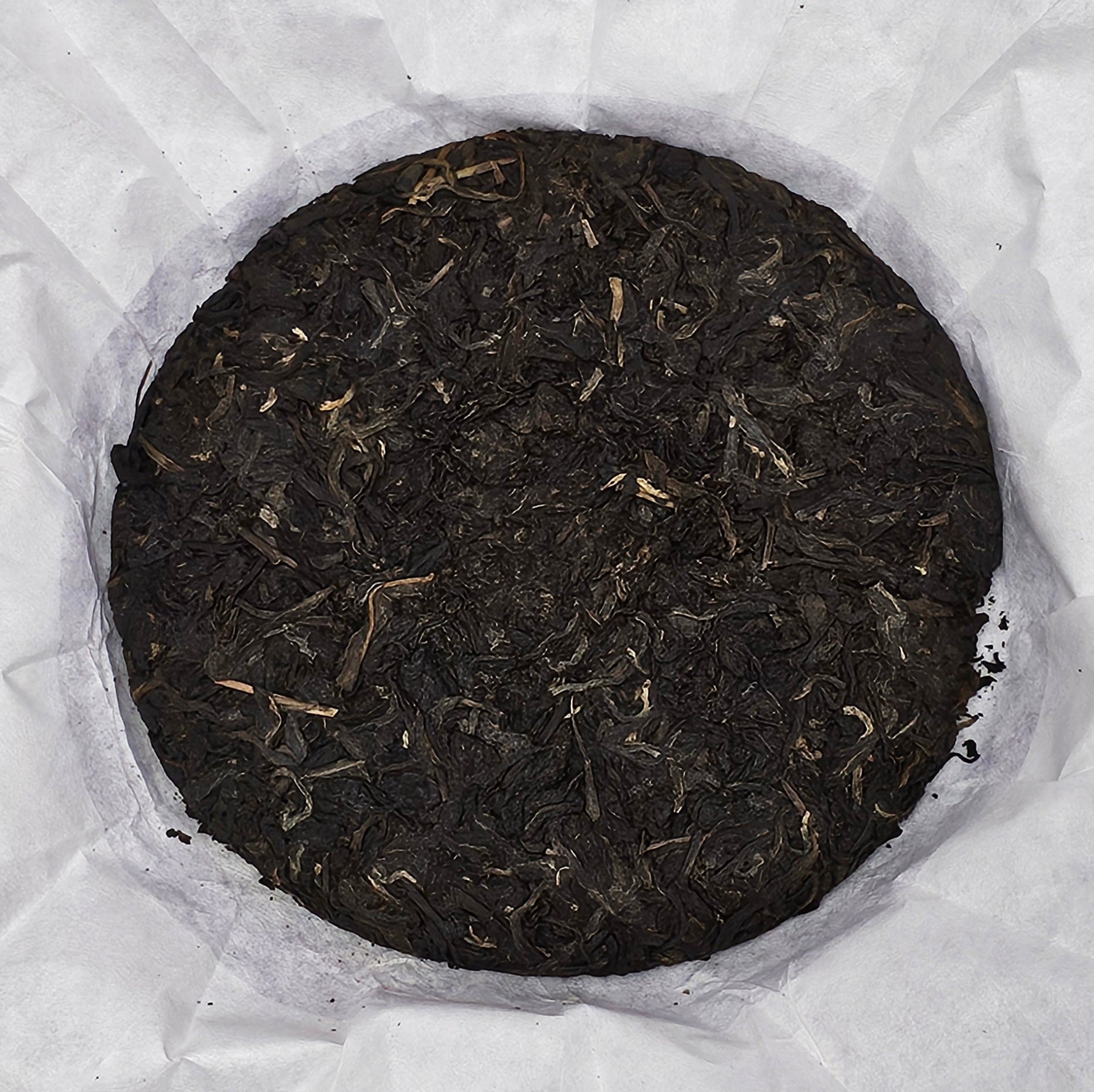 A compressed round cake of dark tea leaves from 2008 Jingmai Mountain (景邁山) Purple Tea (紫茶) 357g, wrapped in white parchment paper, viewed from above by ZenTea Manor-YN003.