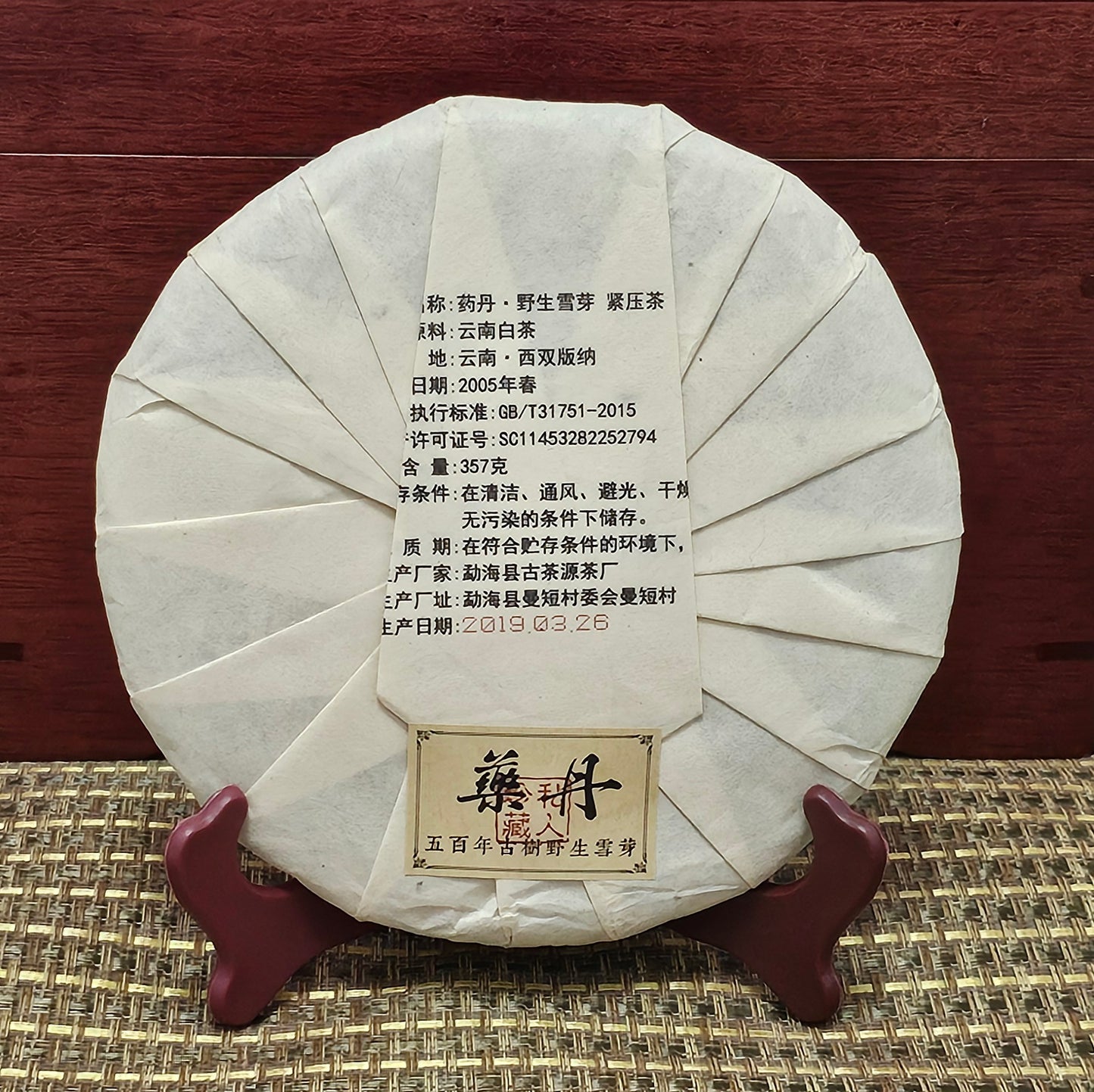 A round, wrapped package of pressed 2005 Wild Ancient Tree Snow Bud Feather White Tea《野生古樹雪芽羽毛白茶》357g from ZenTea Manor-YN003 with labeled information in Chinese characters, displayed on a small wooden stand against a woven surface and red wooden background. This exquisite tea hails from the ancient tea trees of Xishuangbanna.