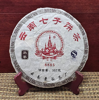 A 2008 Jinggu (景谷) Puerh Ripe Tea cake, labeled "8885" and weighing 357g, with intricate Chinese characters and red graphics, stands elegantly on a wooden stand against a red backdrop, exemplifying the exquisite craftsmanship of ZenTea Manor-YN005.