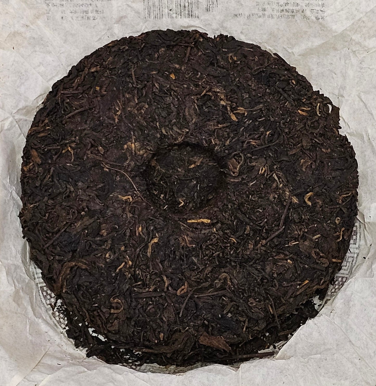 A finely crafted, tightly compressed disc of 2008 Jinggu (景谷) "8885" Puerh Ripe Tea from ZenTea Manor-YN005, weighing 357g, sits on a white crumpled paper background, showcasing its impeccable craftsmanship.