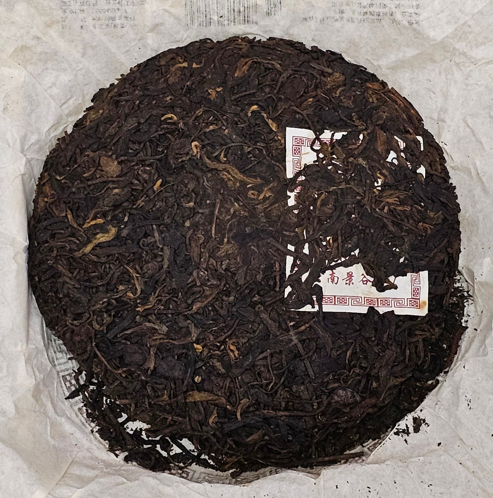 The 2008 Jinggu (景谷) "8885" Puerh Ripe Tea from ZenTea Manor-YN005, weighing 357g, rests as a round disc of dried, dark tea leaves on a white surface, partially covering a piece of paper with printed text and red markings.