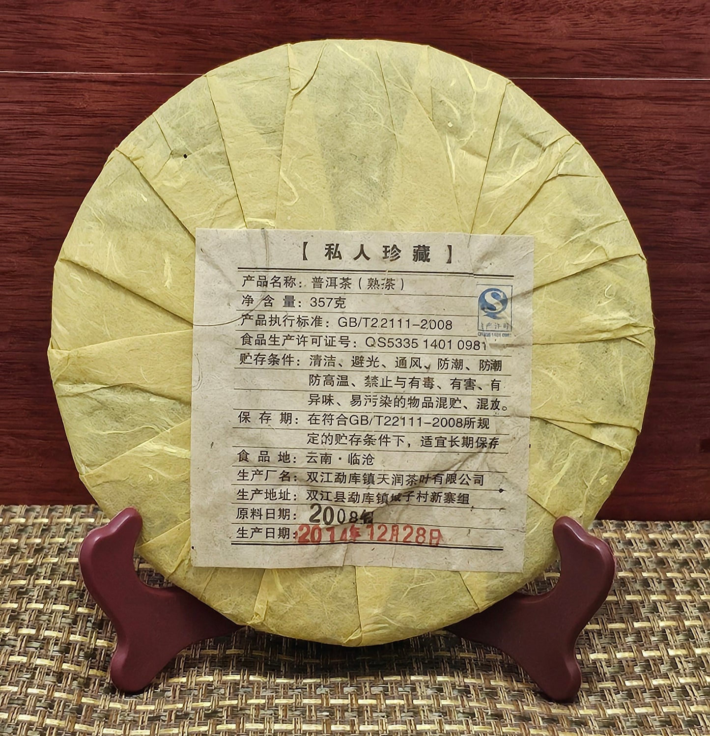 A round, wrapped 2008 Mansong Prince Mountain Tribute Tea ( 曼松王子山貢茶 ) - Wujin No.1 ( 烏金壹號 ) Puerh Ripe Tea 357g by ZenTea Manor-YN005 is displayed on a wooden stand. The wrapper features Chinese text and details, including a manufacturing date of 2008 and a packed date of 2014-12-28.