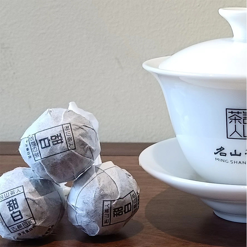 A white lidded tea cup and saucer beside three round, paper-wrapped Sweet White Antique Tree Beads 8g/80g by ZenTea Manor-YN001, all placed on a wooden surface.