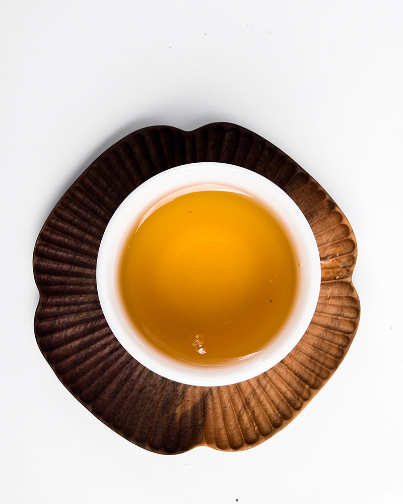 A white bowl containing a yellow liquid that exudes the wild flavor of Yunnan wild tea, placed on a brown, shell-shaped saucer against a white background. The product is Sweet White Antique Tree Beads 8g/80g by ZenTea Manor-YN001.