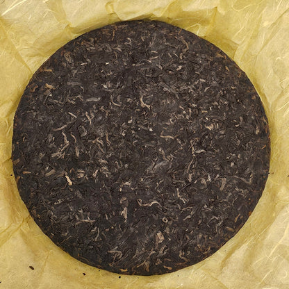 A round, compressed disk of **2008 Mansong Prince Mountain Tribute Tea ( 曼松王子山貢茶 ) - Wujin No.1 ( 烏金壹號 ) Puerh Ripe Tea 357g by ZenTea Manor-YN005** on a yellow crinkled paper.