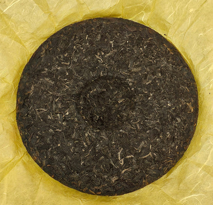 A round disc of compressed 2008 Mansong Prince Mountain Tribute Tea ( 曼松王子山貢茶 ) - Wujin No.1 ( 烏金壹號 ) Puerh Ripe Tea 357g from ZenTea Manor-YN005 sits on yellow paper.