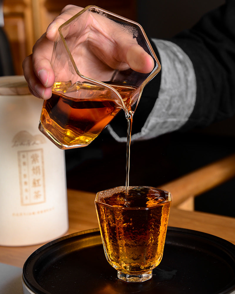 A hand pours a golden-brown Zijuan Black Tea 130g/150g from Zen Tea Master from a geometric glass pitcher into a matching glass on a black tray, showcasing the elegance of this high anthocyanin black tea.