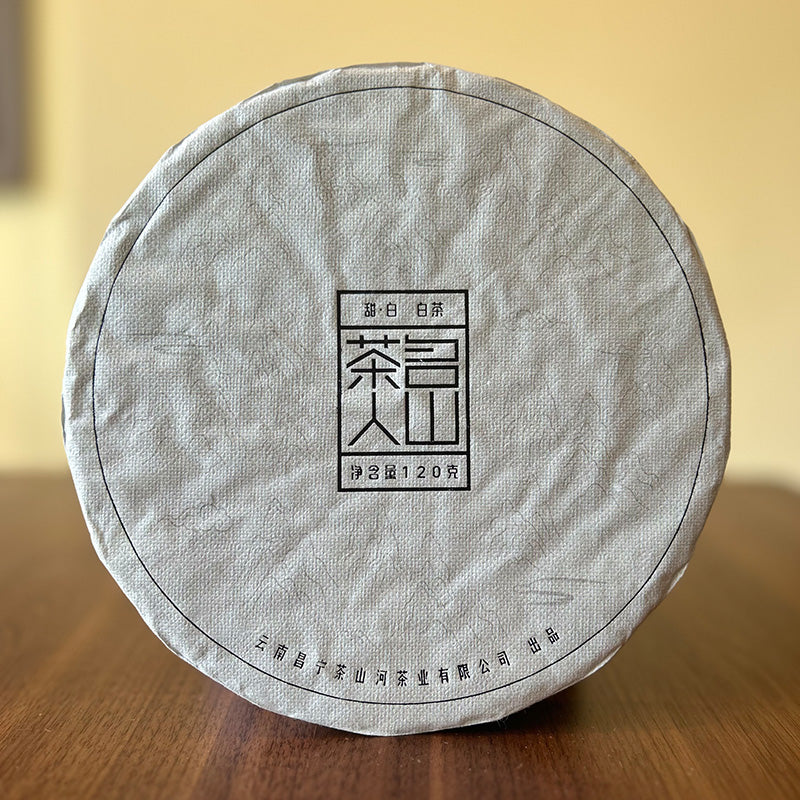 A circular package of Sweet White Black Tea Cake 120g by Zen Tea Master with Chinese characters on it, placed on a wooden surface. The background is blurred.